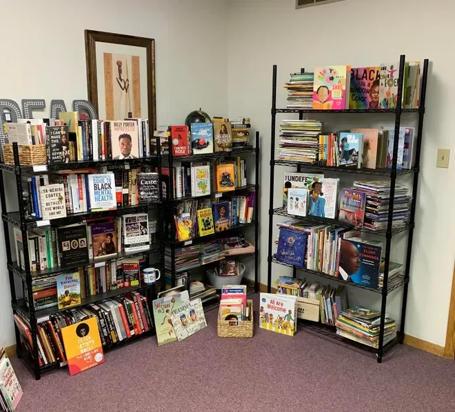 Two black metal bookshelves filled with a variety of books, some books leaning and stacked, with additional books and signs on the floor in front of them, in a room with beige walls and purple carpet.