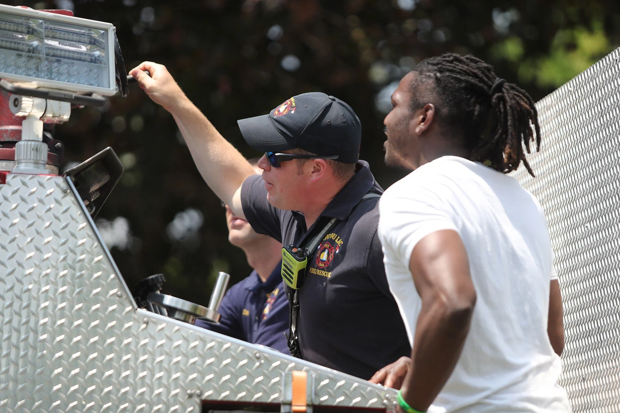 Two firefighters and a man with dreadlocks standing by a fire truck, with one firefighter adjusting equipment.