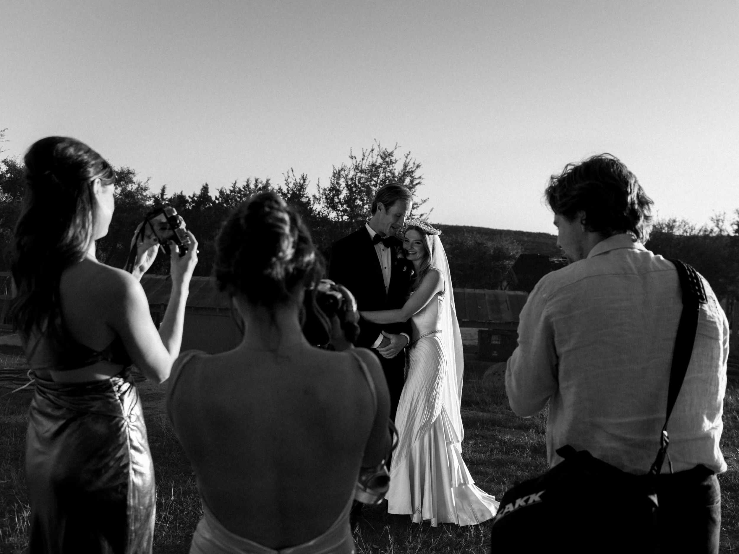 A black and white photograph of a wedding scene outdoors during sunset, with a bride and groom embracing and smiling, surrounded by friends and photographers capturing the moment.