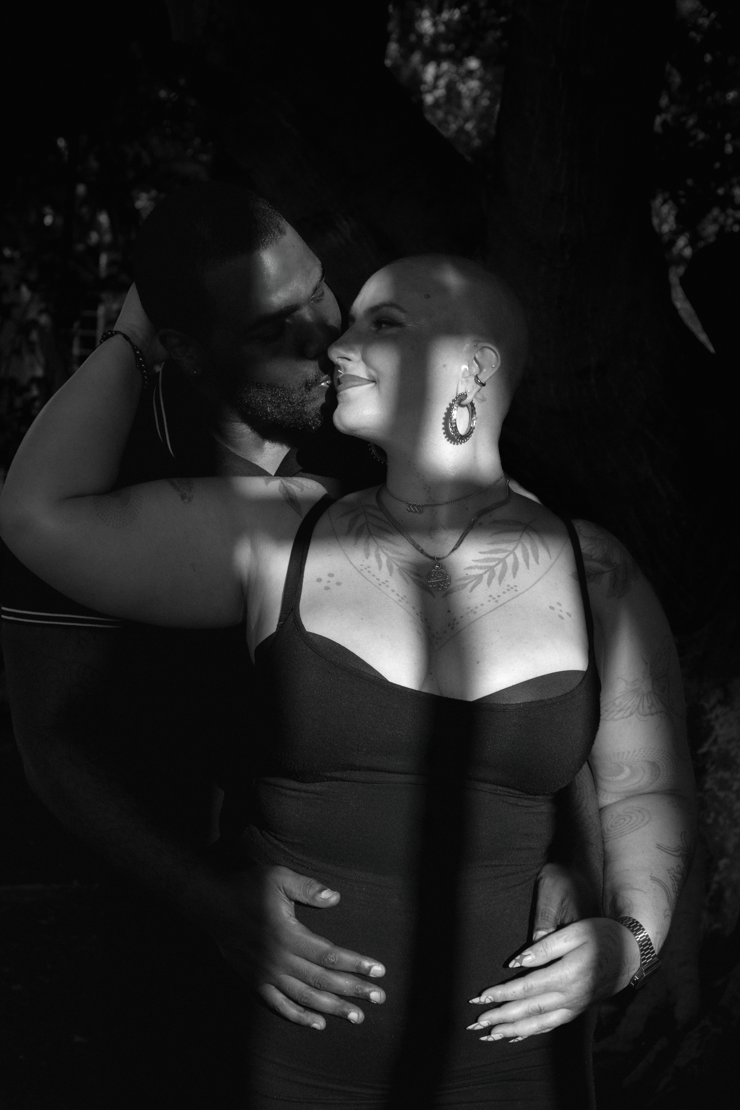 A couple sharing an intimate moment outdoors at night, with the man leaning in to kiss the woman, who has a tattooed upper chest, multiple earrings, and short hair, illuminated by a narrow beam of light.