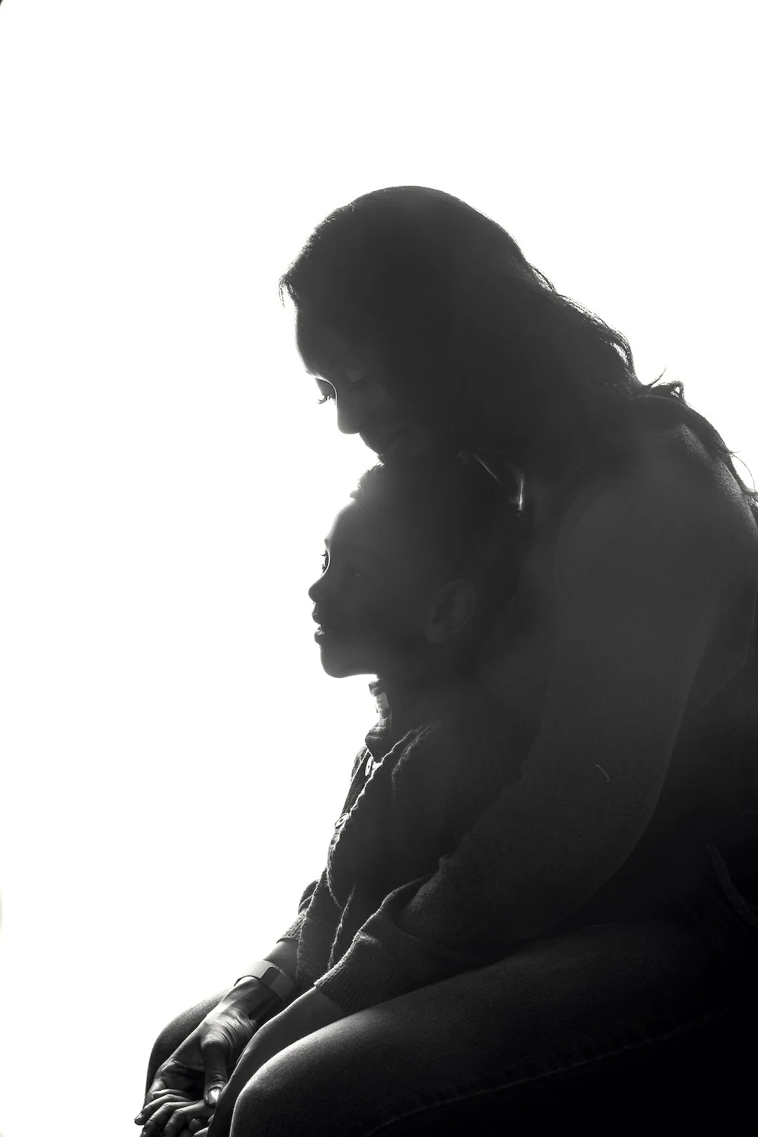 Silhouette of a woman embracing a child, both sitting on the floor with the woman leaning her head against the child's head.