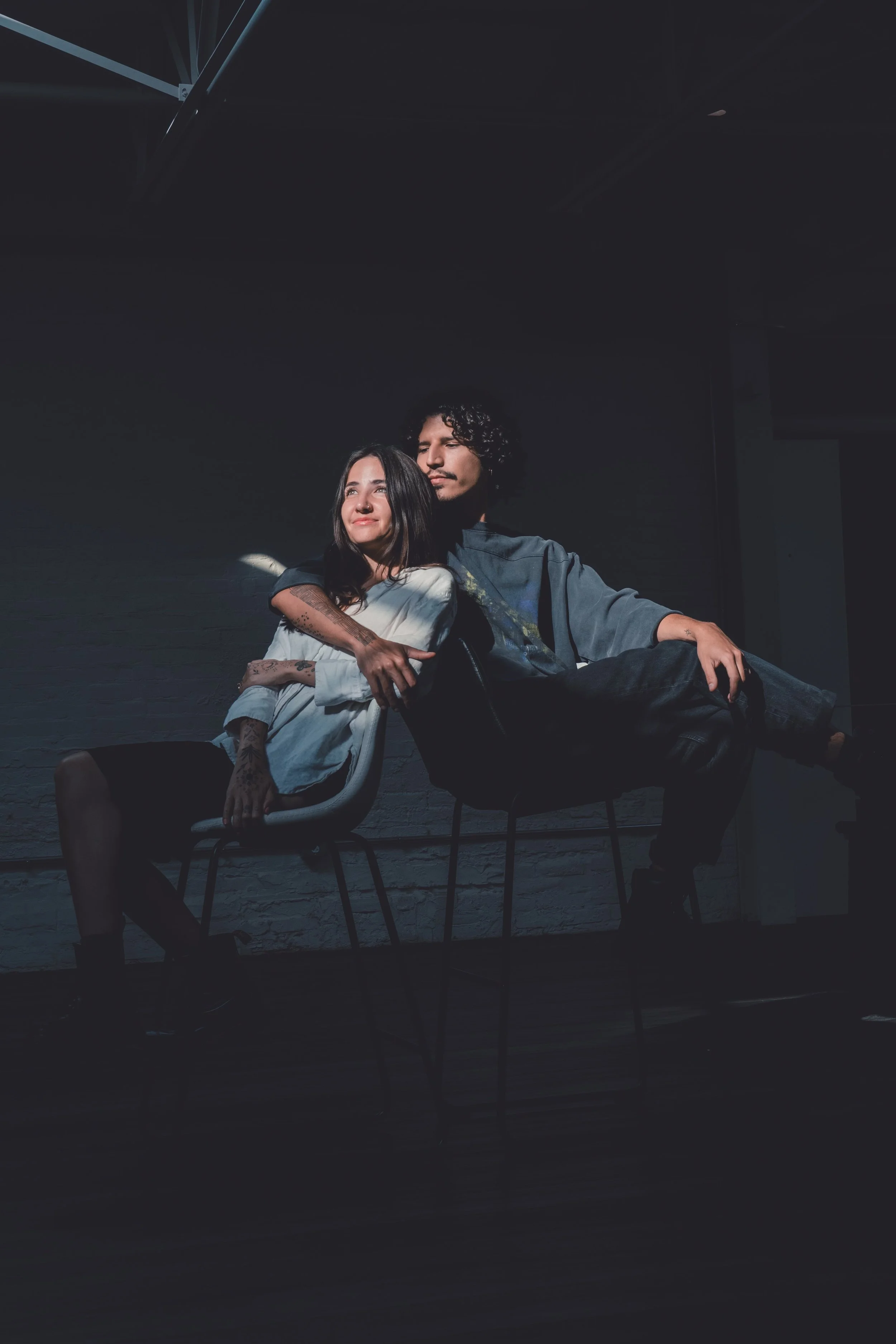 A man and woman sit on chairs in a dark room, with the woman leaning against the man, both appearing relaxed and content.