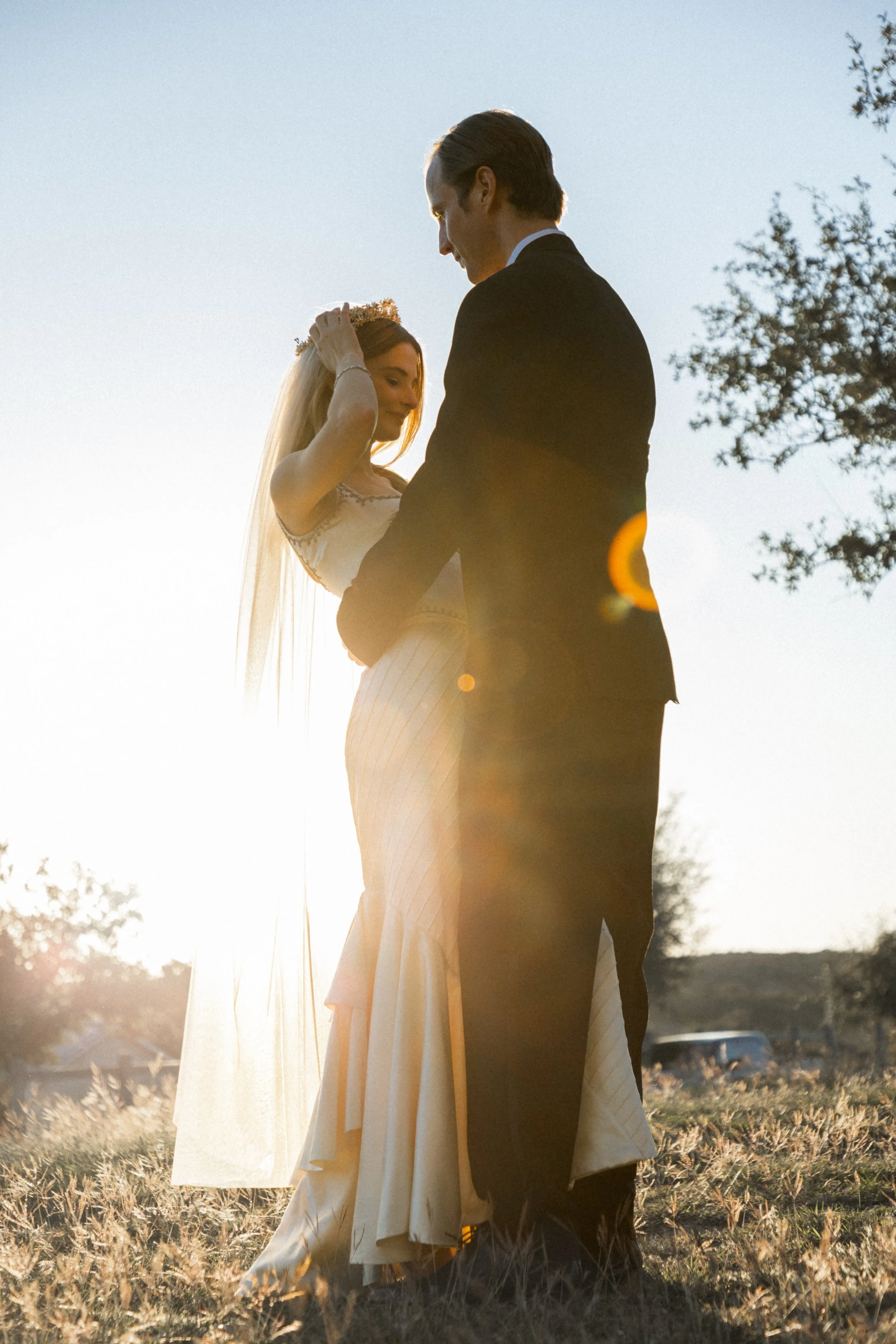 A bride and groom standing outdoors during sunset, holding hands and gazing at each other, with sunlight creating a warm glow.
