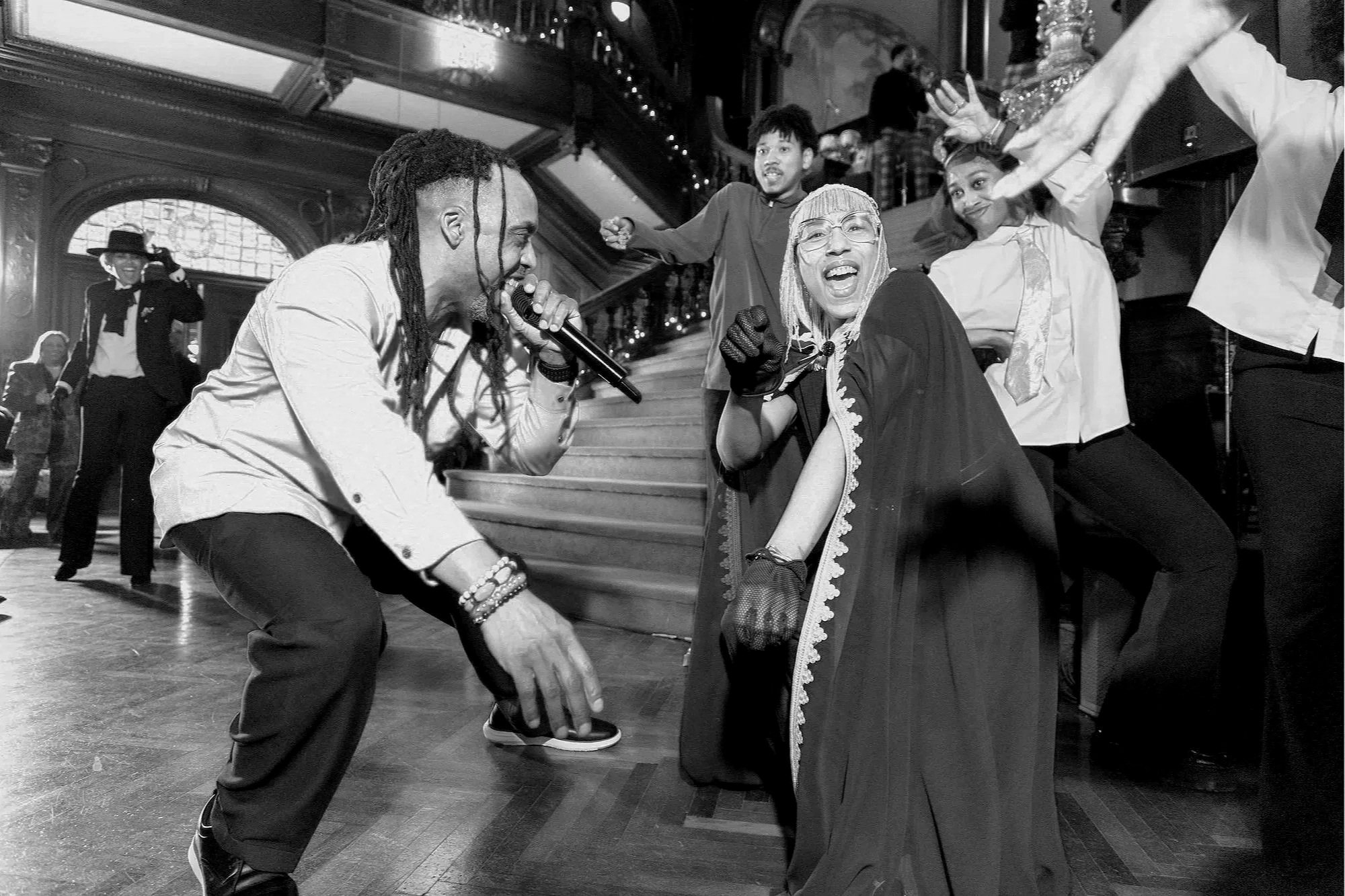 People dancing and singing at a lively party or celebration in an elegant interior, with a man holding a microphone, and others smiling and dancing around him.