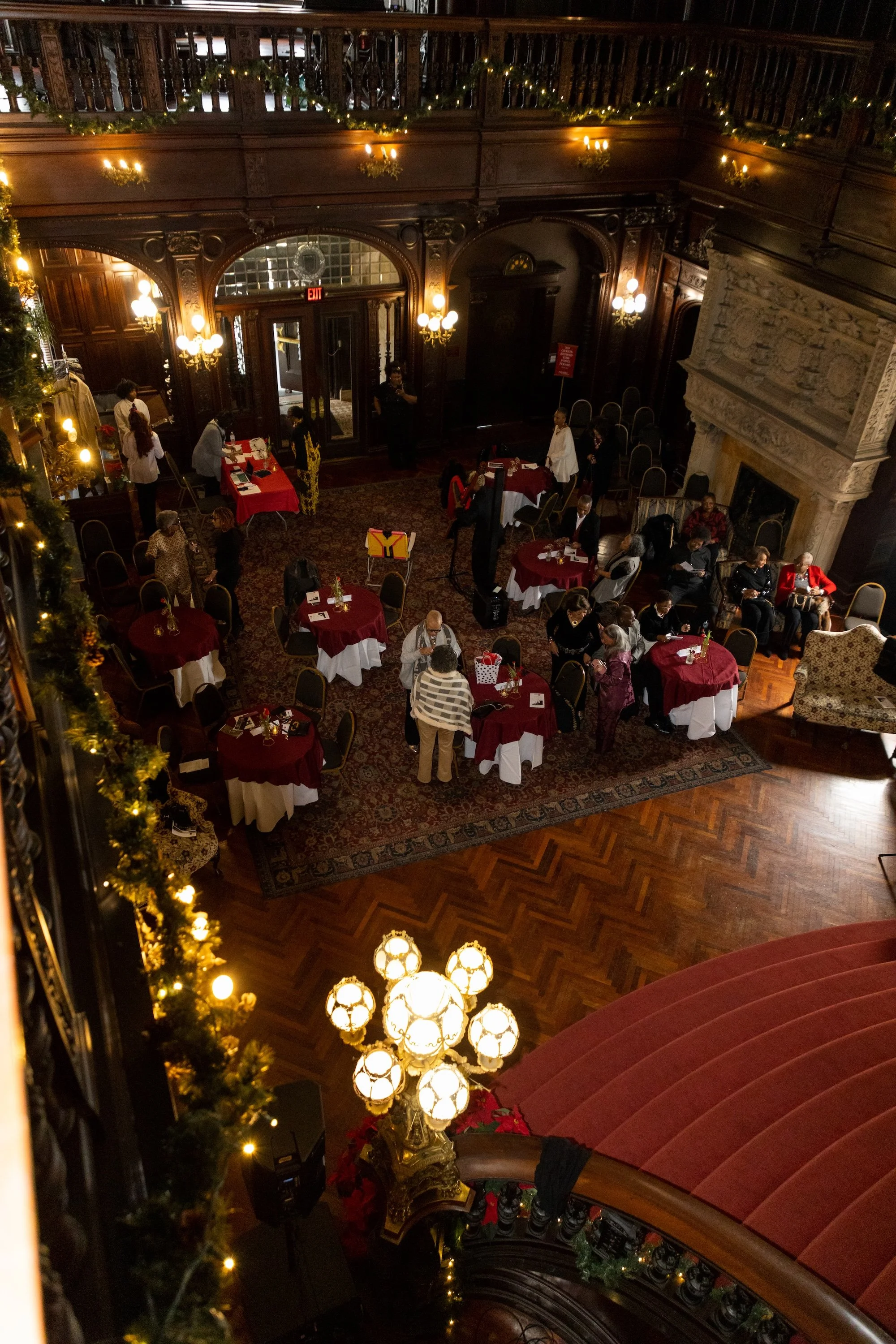 An elegant, festive indoor event with people sitting at round tables with red and white tablecloths, decorated with holiday decorations. The room features ornate woodwork, a fireplace, and warm lighting, with Christmas greenery and lights as holiday decor.