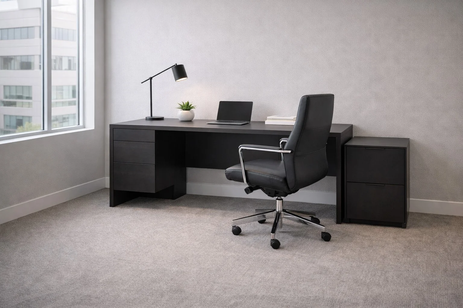 An empty office with a black desk, black rolling chair, a small storage cabinet, a lamp, a potted plant, and a laptop on the desk, with a window showing a cityscape outside.