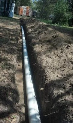 A long trench with a large pipe installed inside, surrounded by dirt, with trees and a shed in the background.