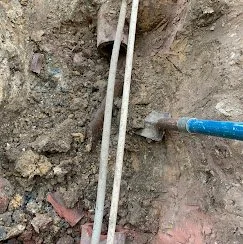 Close-up of a small trench with two white pipes and a blue-handled trowel in soil.