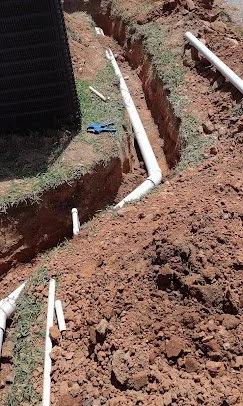 A construction site showing a trench with white PVC pipes installed for plumbing, with a black screen or barrier on the left side.