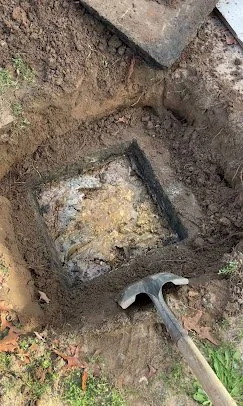 Excavation site with a rectangular hole in the ground containing concrete debris, surrounded by dirt and a hammer placed nearby.