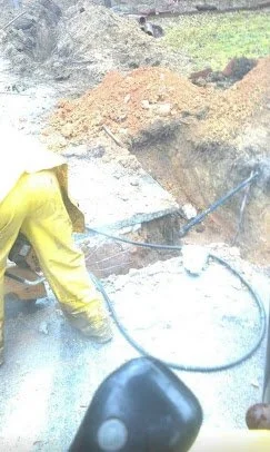 Person in yellow jacket using a hose to spray water on a muddy area outdoors.