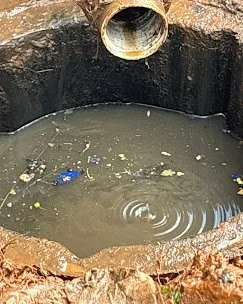 A rusty pipe leading into a stagnant, muddy water-filled hole with debris and trash.