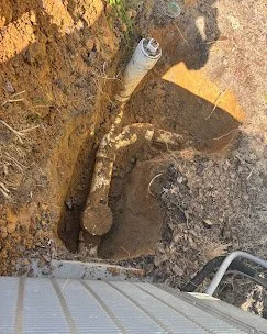 A snapshot of an underground plumbing repair, showing a pipe within a dug trench in the ground, with a pipe fitting visible.