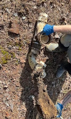 Person excavating dirt to uncover an old, rusted pipe in a muddy outdoor environment.