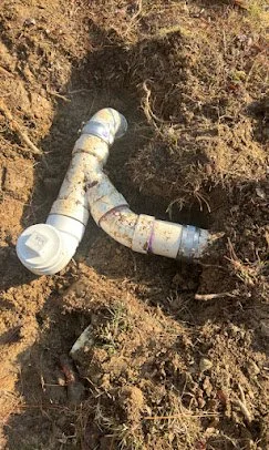 A broken white PVC pipe lying in a dug hole in the ground.