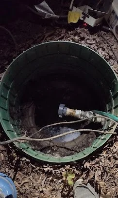 A partially buried green plant pot with a hose and some wires inside, on dirt ground.