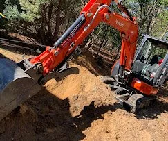 An orange mini excavator digging into the earth outdoors, with trees in the background.