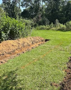 A backyard with freshly dug soil along the edge of a grassy lawn, surrounded by trees and bushes.
