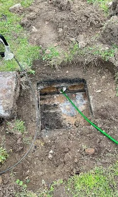 An open rectangular underground utility access or valve box in a dirt yard, with a green hose connected inside and a water hose spraying water into it.