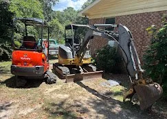 Small orange excavator with a black arm and bucket, parked on a lawn next to a house, with trees and bushes in the background.