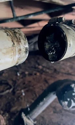 Close-up of a dirty pipe fitting connected to a pipe with a black cap, with wooden and metal structures in the background.