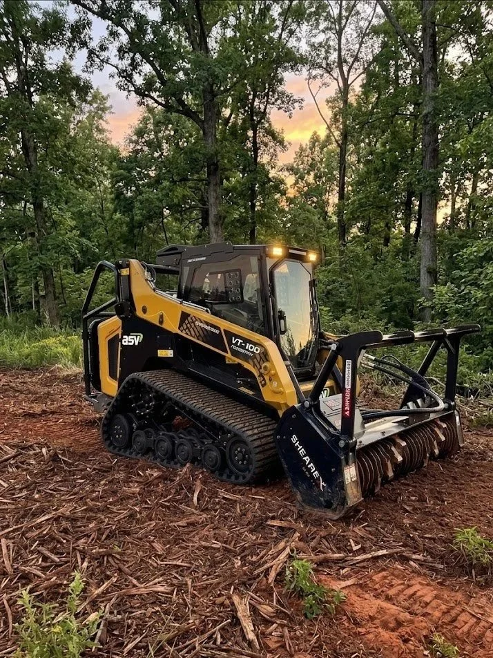 A yellow and black compact track loader with a shearing attachment on the front, operating in a cleared wooded area.