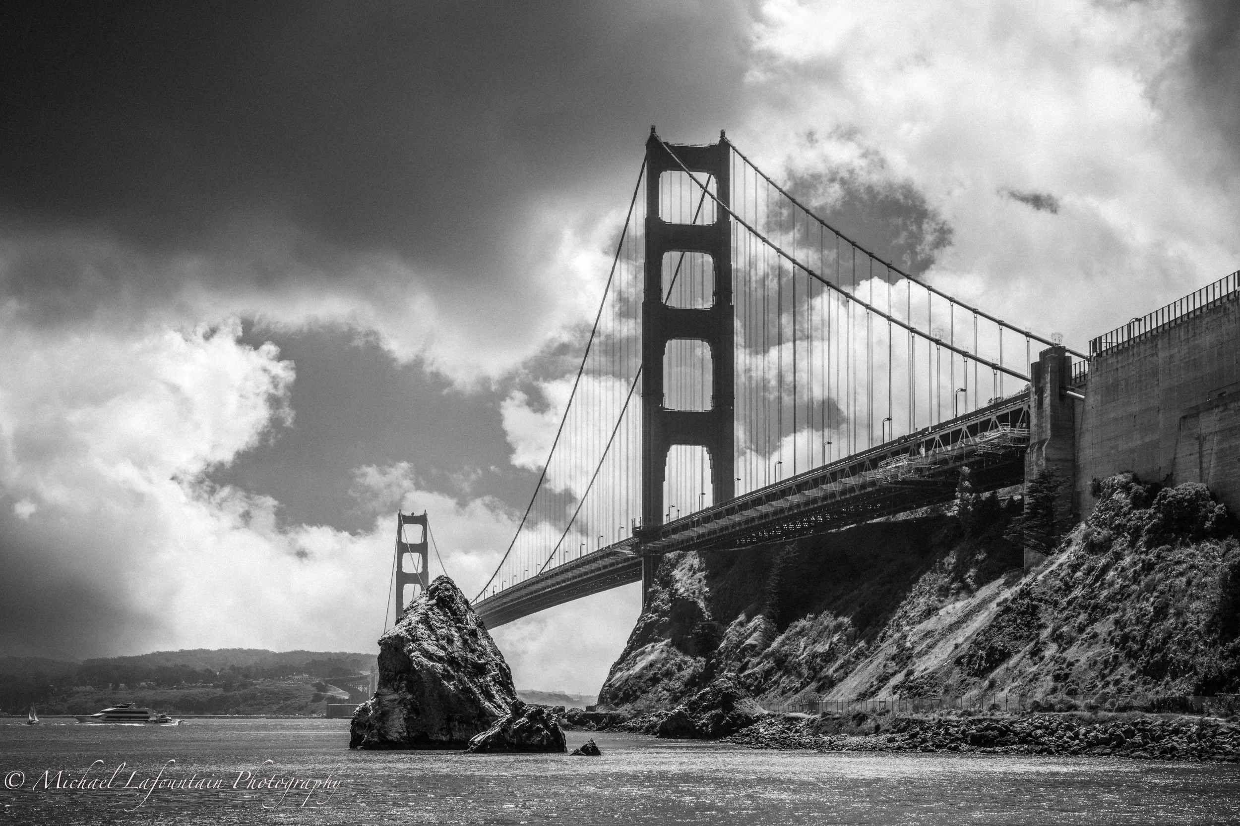 Black and white photo of the Golden Gate Bridge in San Francisco, with large rocks in the water in the foreground and cloudy sky above.