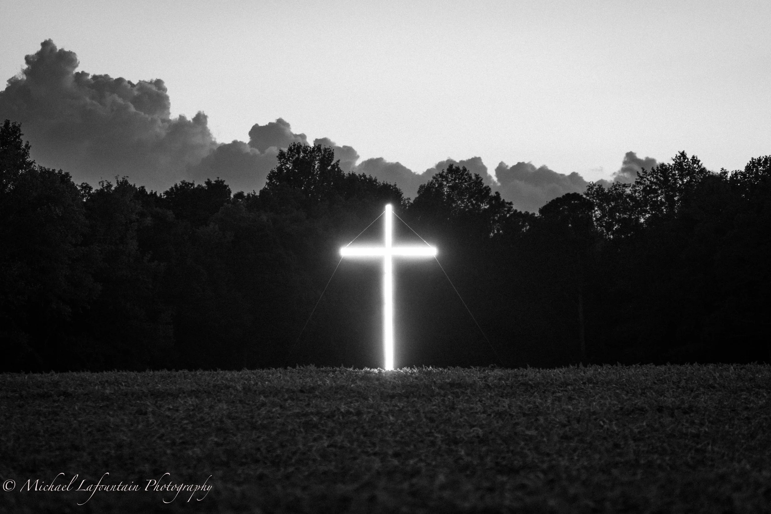 A glowing cross illuminated at night, mounted on a hill with a background of trees and clouds.