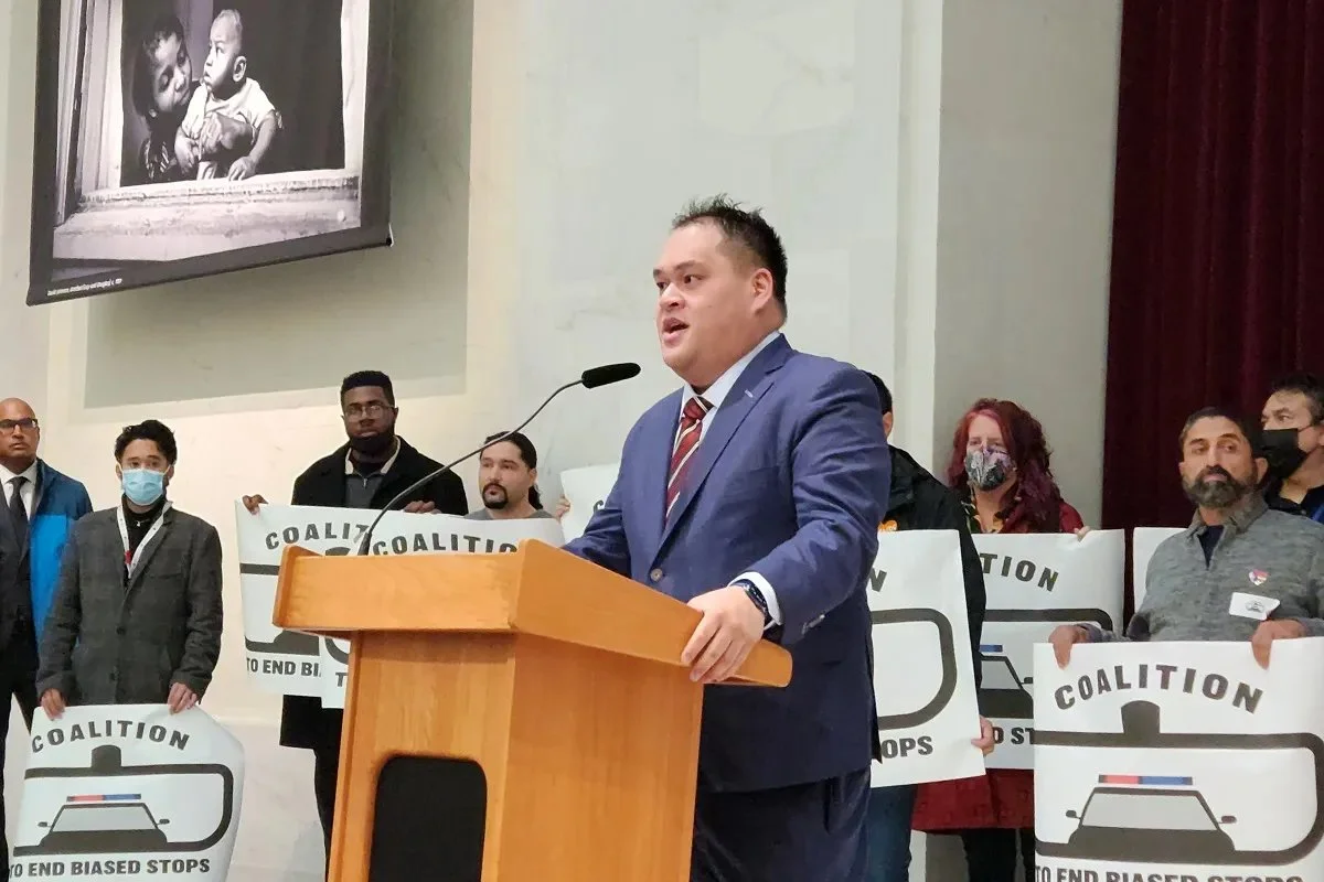 Man in a blue suit speaking at a podium during a protest or rally, with several people holding signs that read 'Coalition to End Biased Stops,' in a large indoor space.