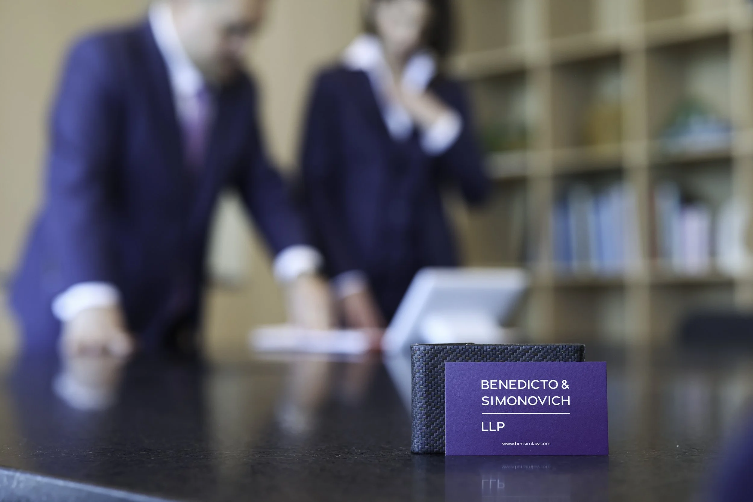 Business card for Benedict & Simonovich LLP placed on a dark conference table with two blurred people in business attire in the background