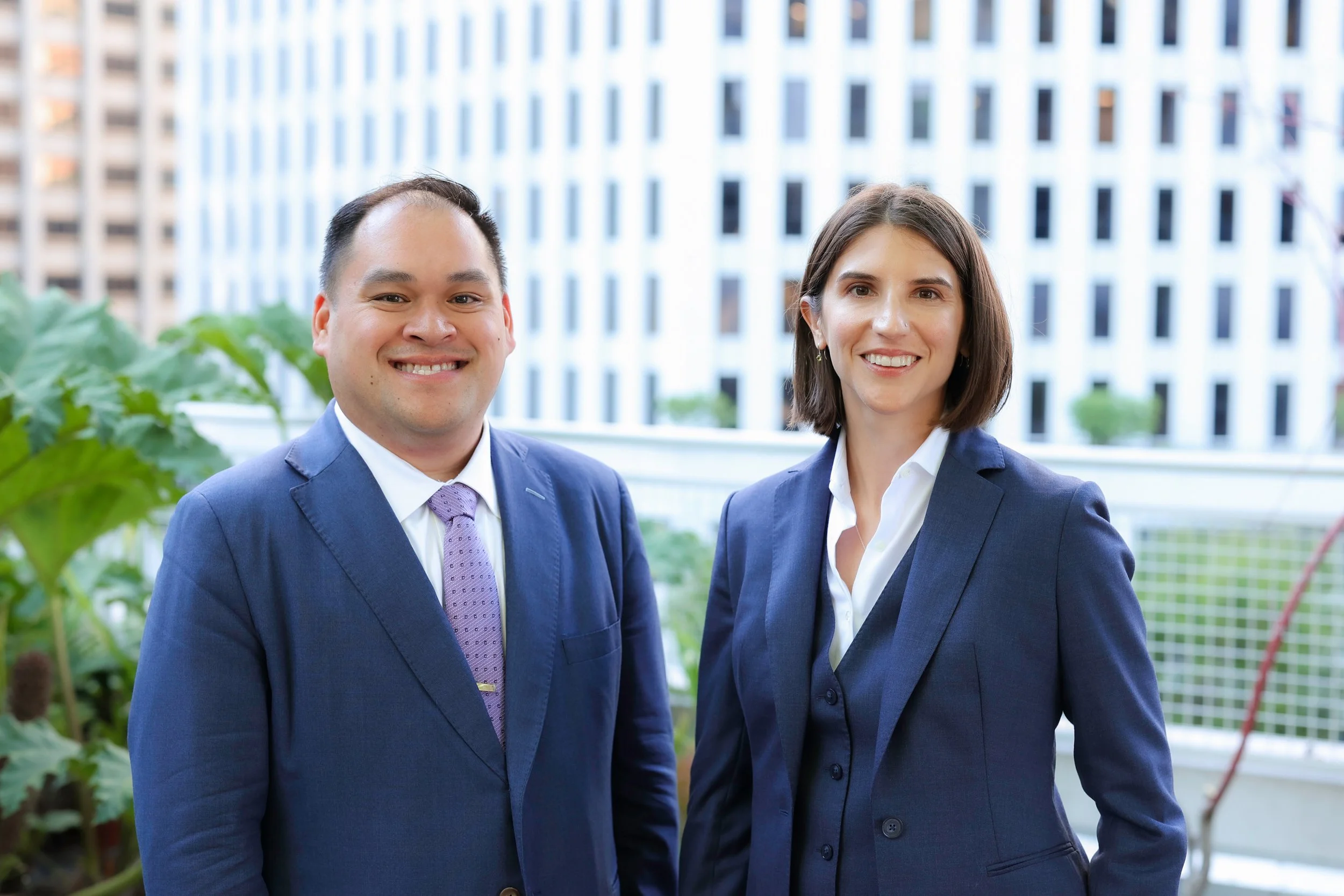 Two professionally dressed individuals smiling on a balcony with city buildings and green plants in the background.