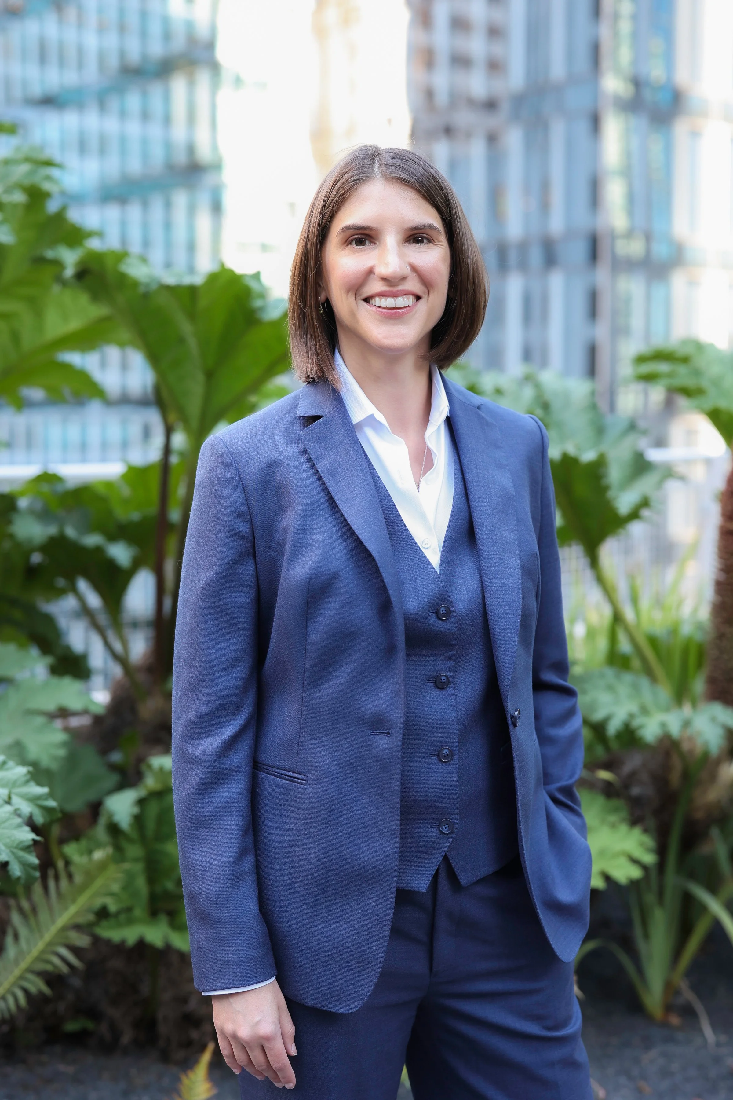 A woman in a blue business suit standing outdoors in a cityscape with greenery and tall buildings in the background.