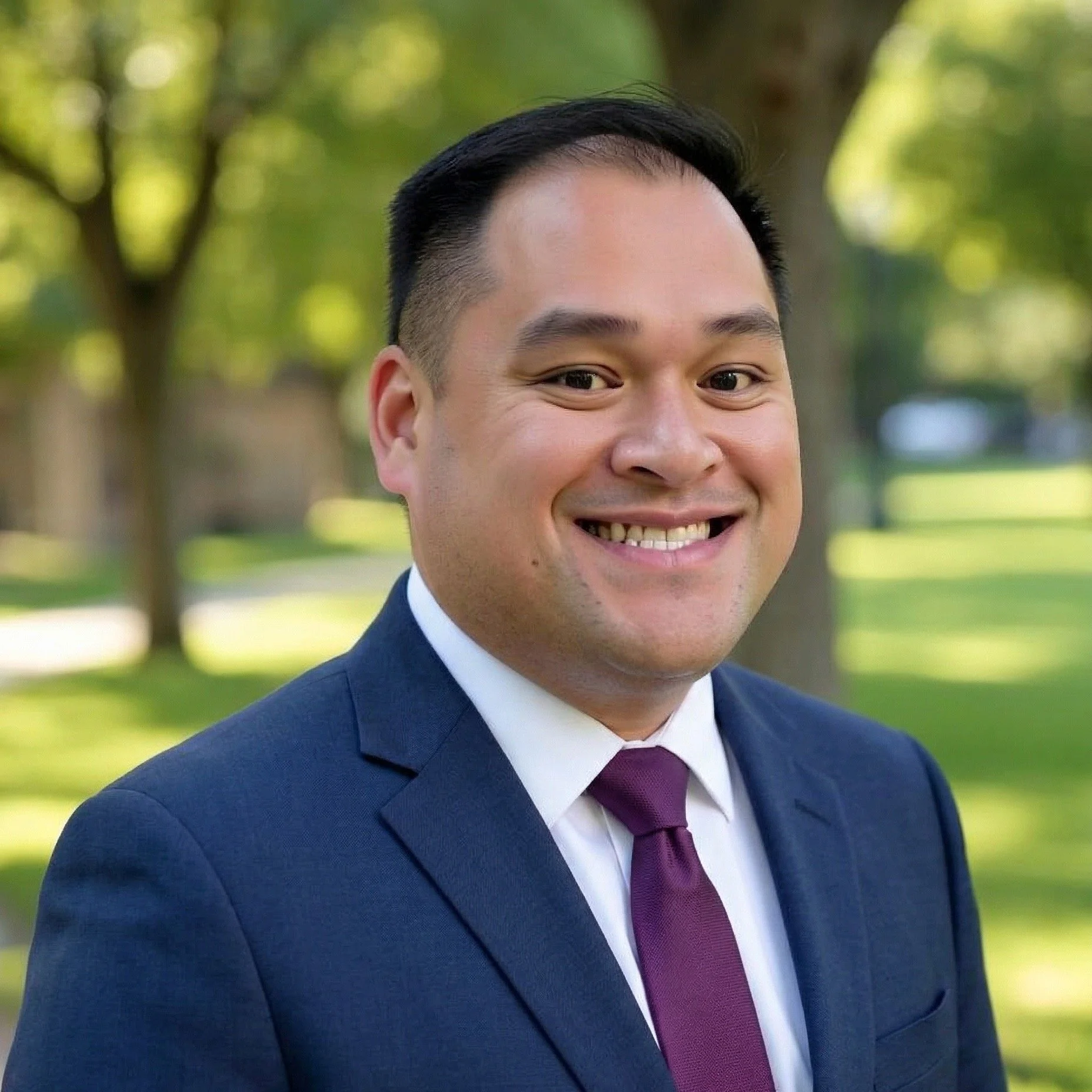 A photo of BenSim Law founding partner Kevin Benedicto. A man in a navy blue suit, white shirt, and maroon tie smiling outdoors in a park with green trees in the background.
