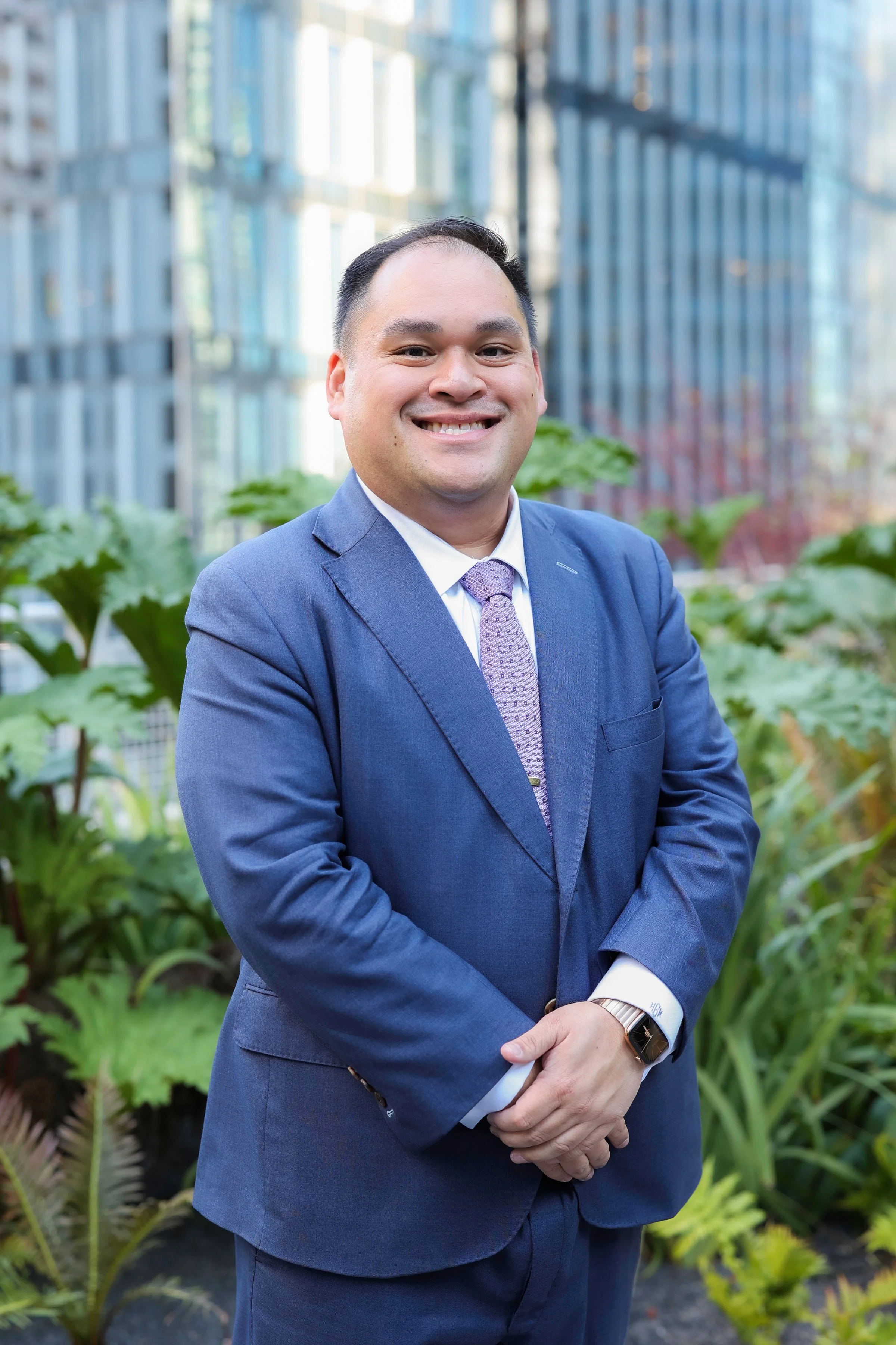 A smiling man dressed in a blue suit, white shirt, and purple tie standing outdoors with green plants and modern glass buildings in the background.