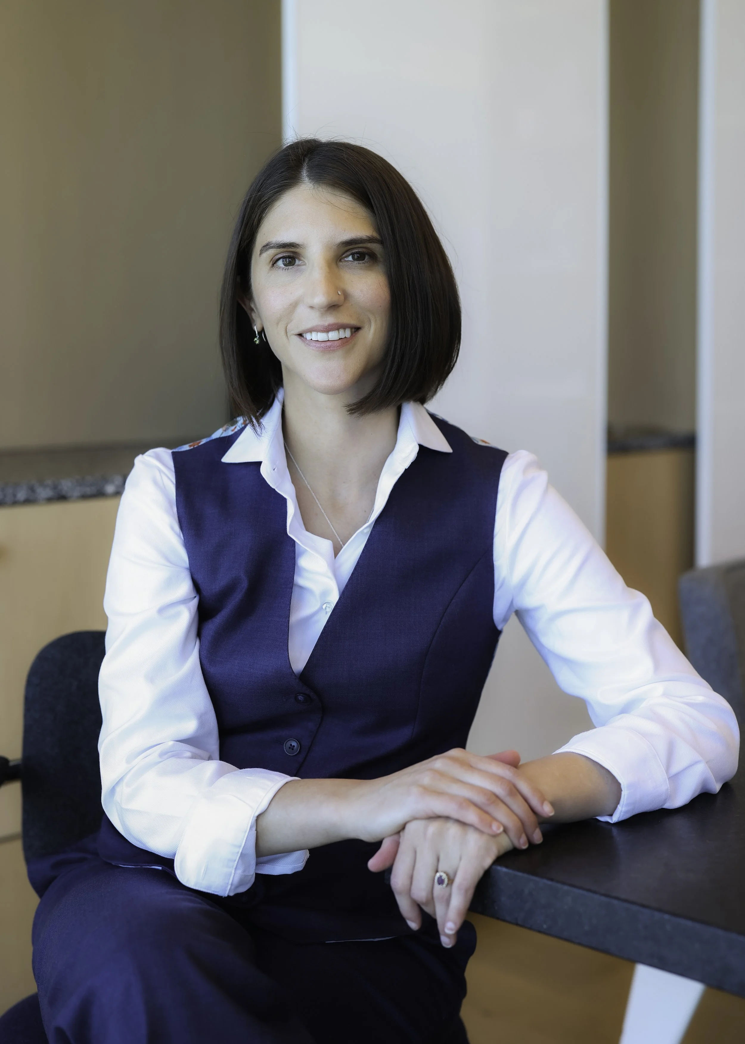 A woman with shoulder-length dark hair smiling, dressed in a white shirt and navy blue vest, sitting at a conference table in an office.