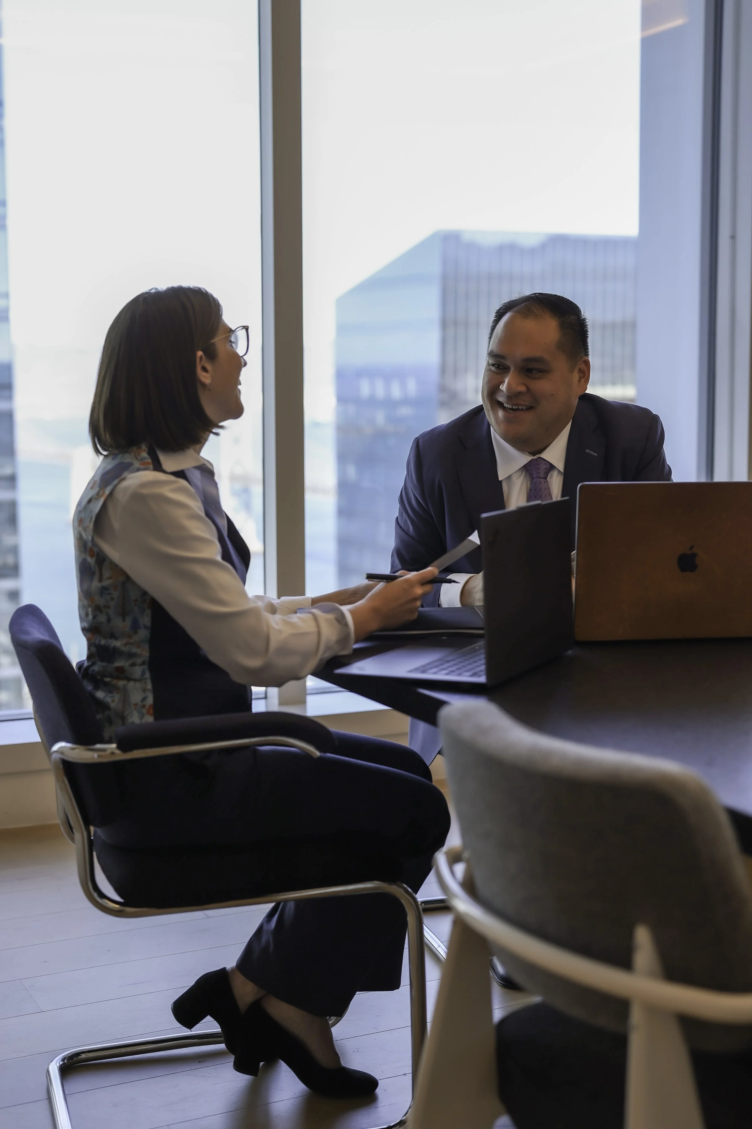 Two business professionals, a man and a woman, are sitting at a table in a modern office with large windows and city buildings outside. They are smiling and engaging in conversation, with laptops in front of them.