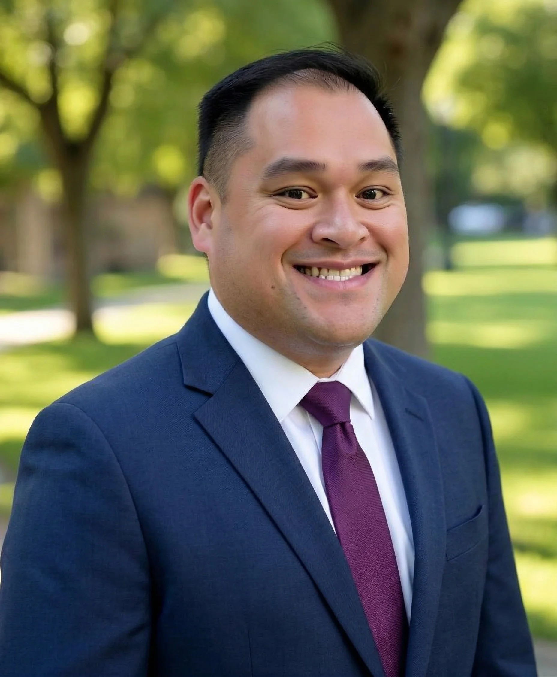 A photo of BenSim Law founding partner Kevin Benedicto. A man in a navy blue suit, white shirt, and maroon tie smiling outdoors in a park with green trees in the background.