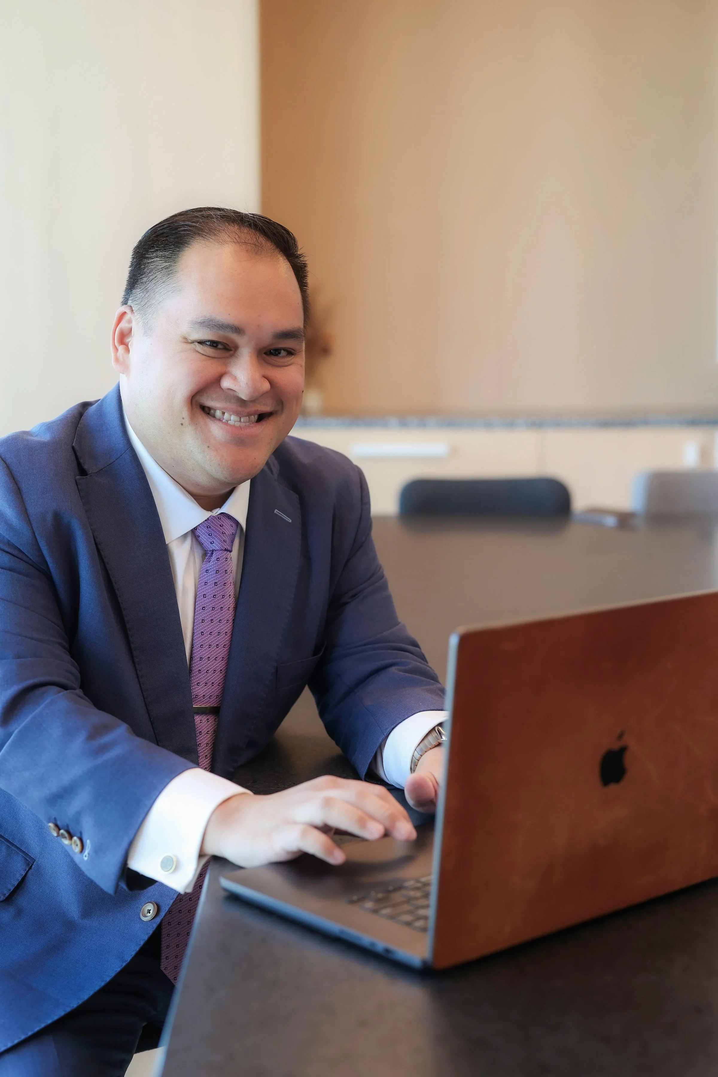 A smiling man in a suit working on a laptop in a conference room.