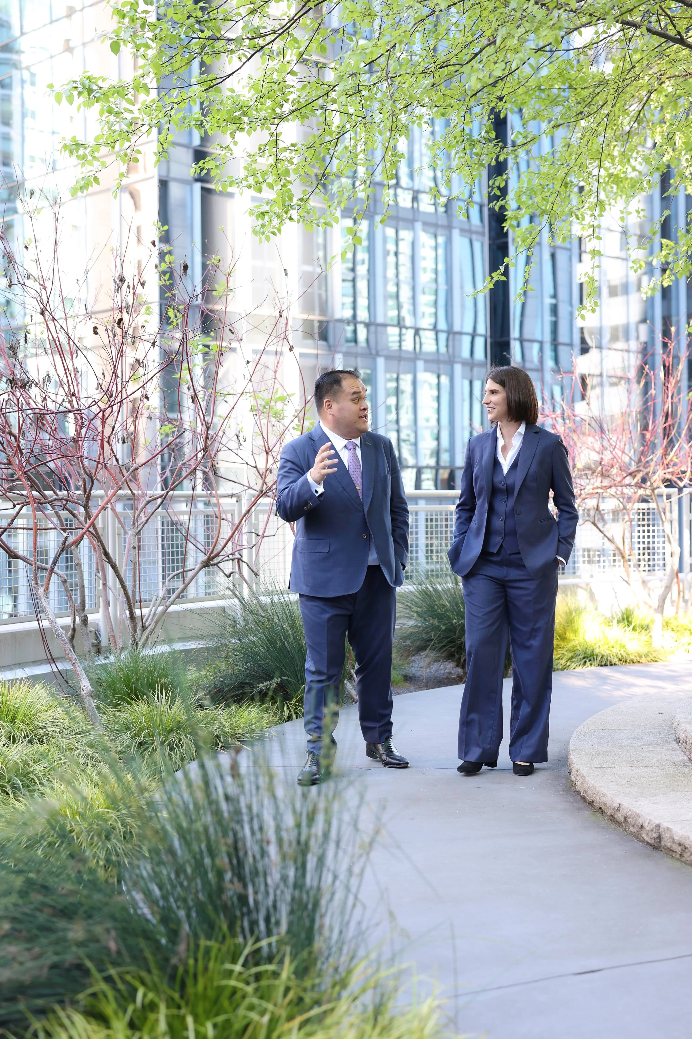 Two businesspeople, a man and a woman, in formal suits, talking outside on a city sidewalk with modern glass buildings and trees around them.