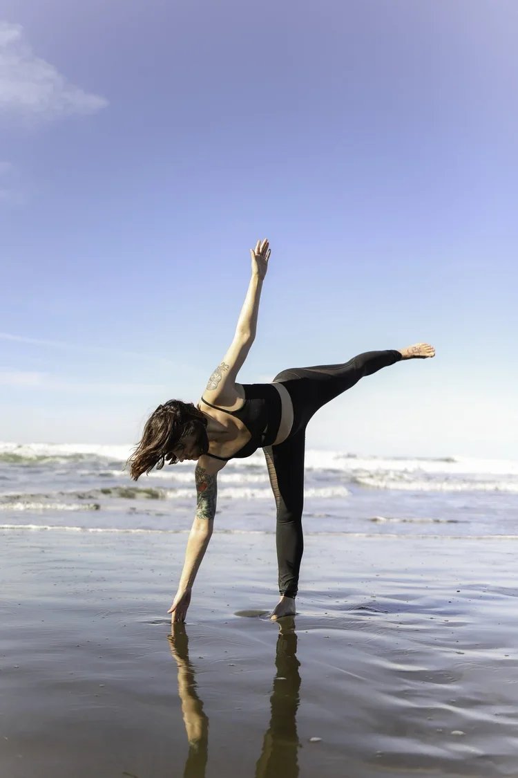 A woman in black athletic clothing practicing yoga on the beach near the water, balancing on one hand with her other hand and opposite leg extended outward.