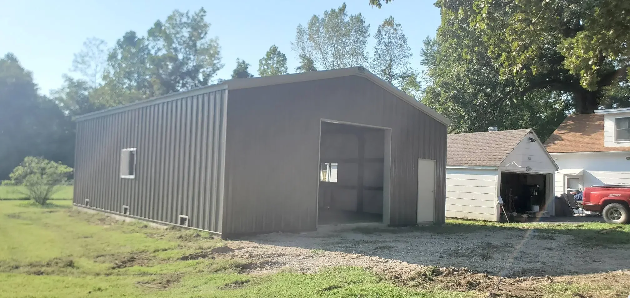 A metal storage shed under construction in a residential backyard with a house, trees, a red truck, and lawn chairs nearby.