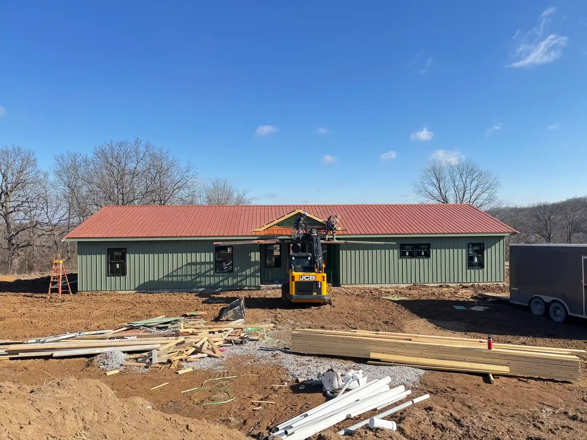 Construction site with a green building, red metal roof, construction worker on a crane installing the roof, wooden planks, and construction materials on dirt ground, with a trailer and leafless trees in the background under a blue sky.