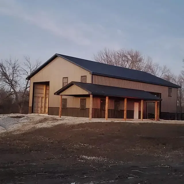 A two-story barn with metal siding, large doors, and a porch supported by wooden posts, situated on a dirt lot with leafless trees in the background.