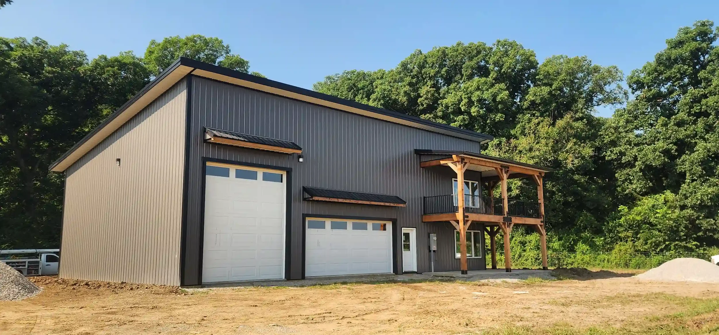 New modern two-story house with metal siding, white garage doors, wooden balcony, and surrounded by green trees.
