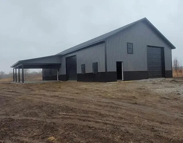 Large metal warehouse or barn with a sloped roof, black garage doors, and small windows, situated on a dirt lot in a rural area under an overcast sky.