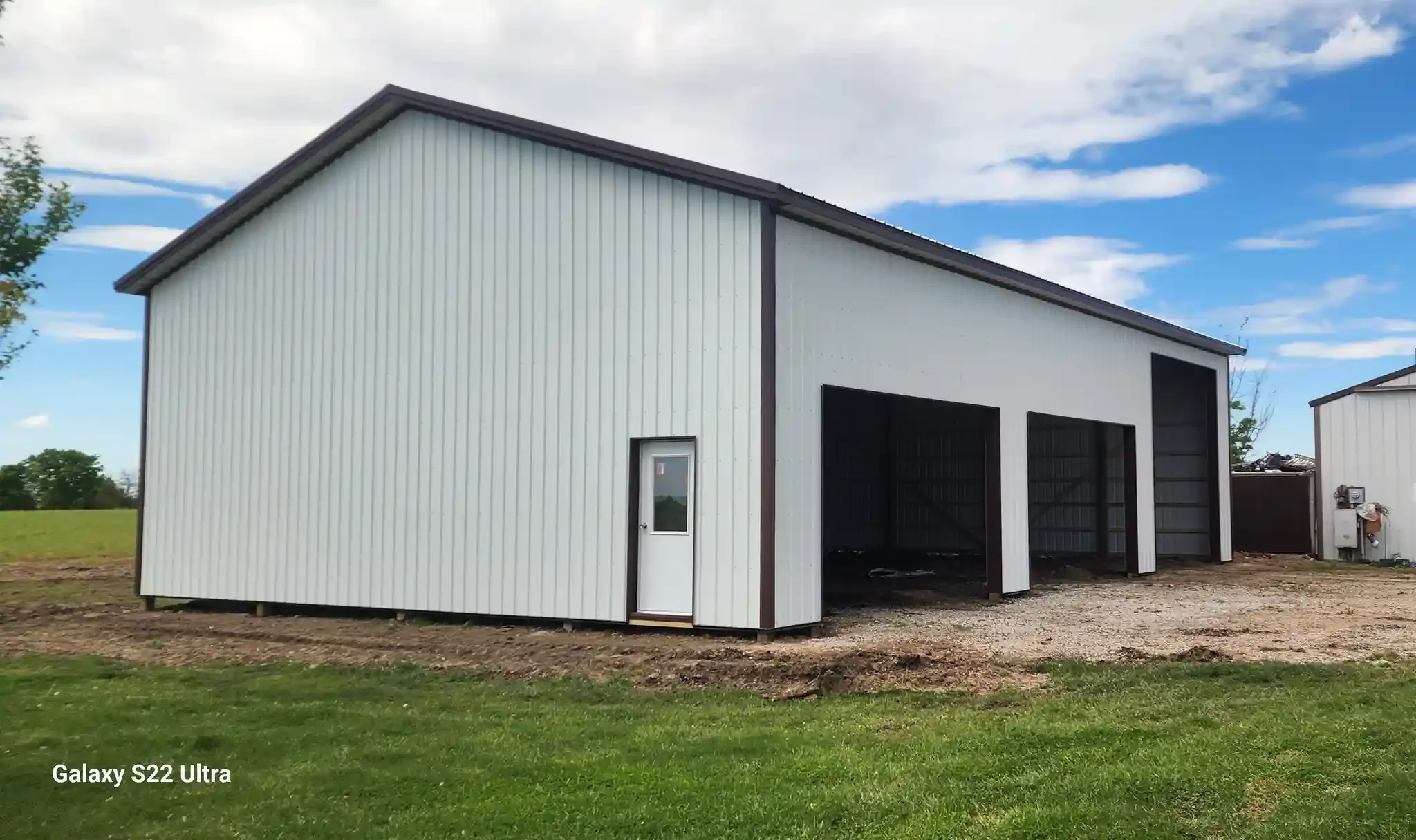 A large white metal building with three garage doors and a small white door on the side, situated on a grassy area with patches of dirt and a partly cloudy sky above.