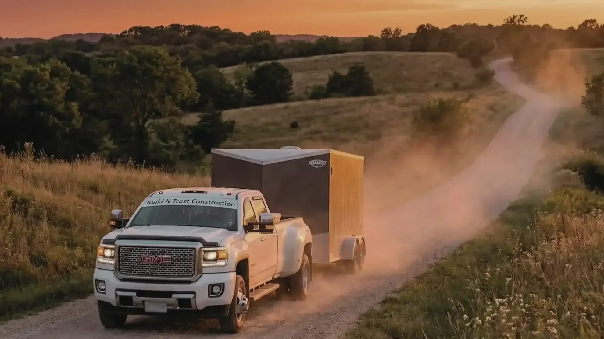 A white GMC truck pulling a large black enclosed trailer on a dirt road in a rural area with grassy fields and trees, with a sunset sky in the background.