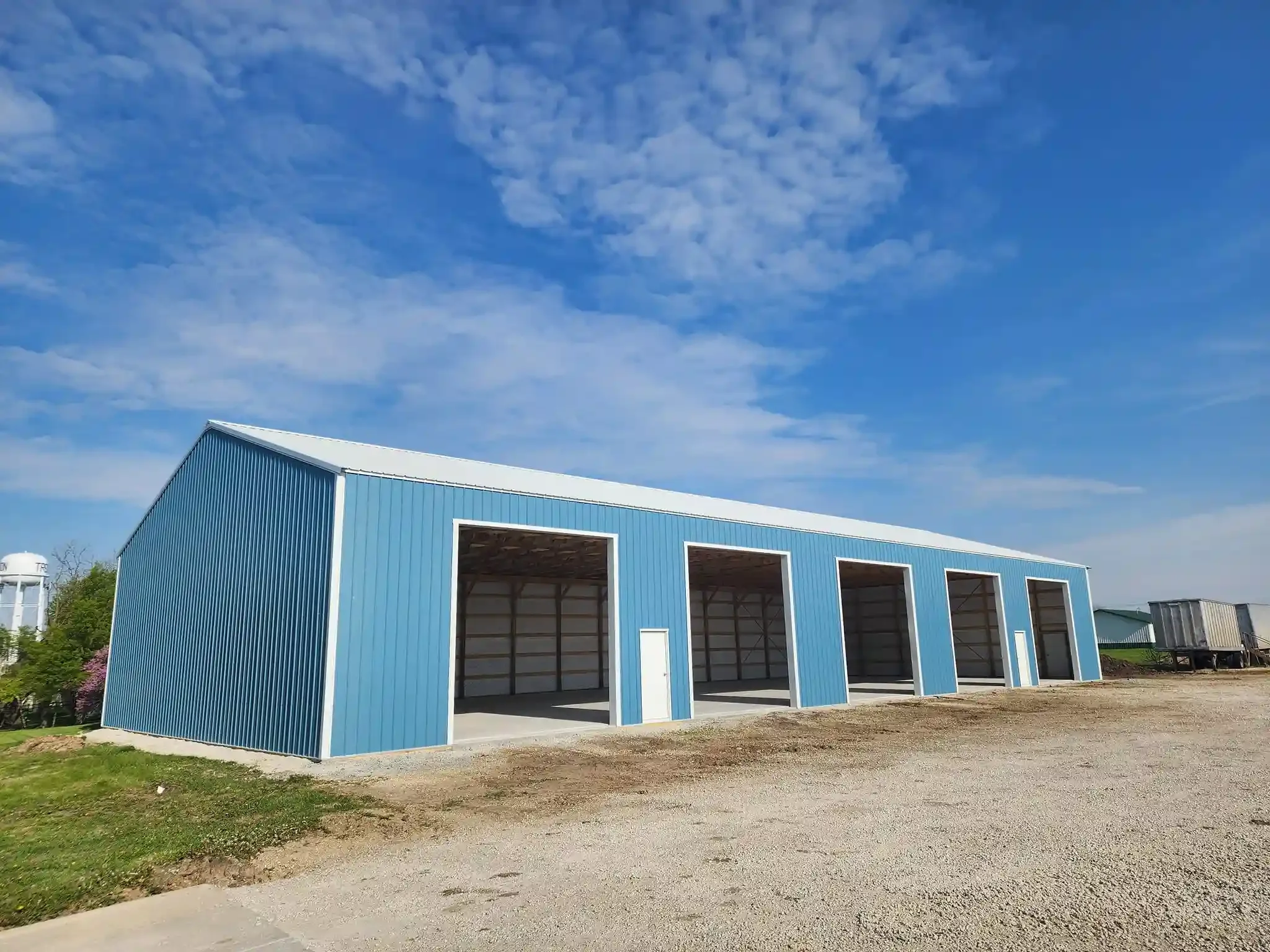 A large blue metal building with four open bays, situated on a gravel lot with some grass, under a partly cloudy blue sky.