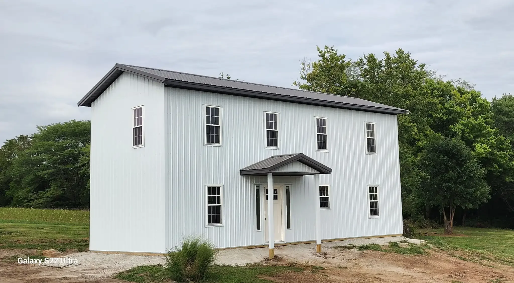 White two-story house with black roof and multiple windows, surrounded by green trees and a grassy yard.