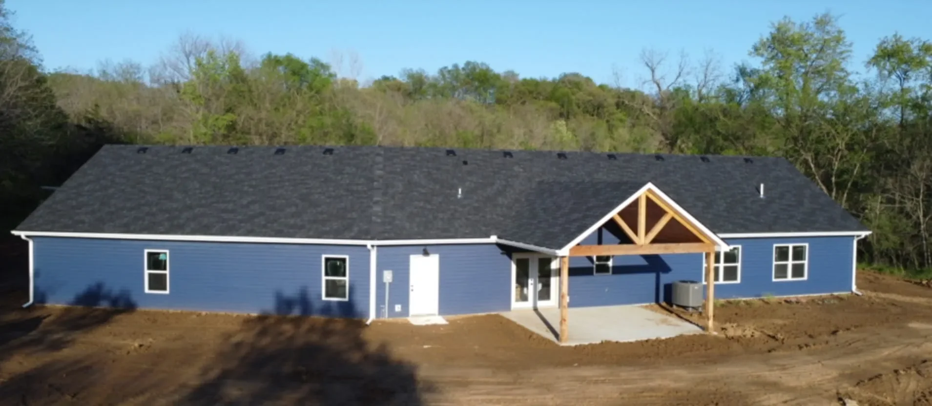 New blue house with black roof and white trim, wooden porch framing entrance, surrounded by bare dirt yard, with trees and a clear blue sky in background.